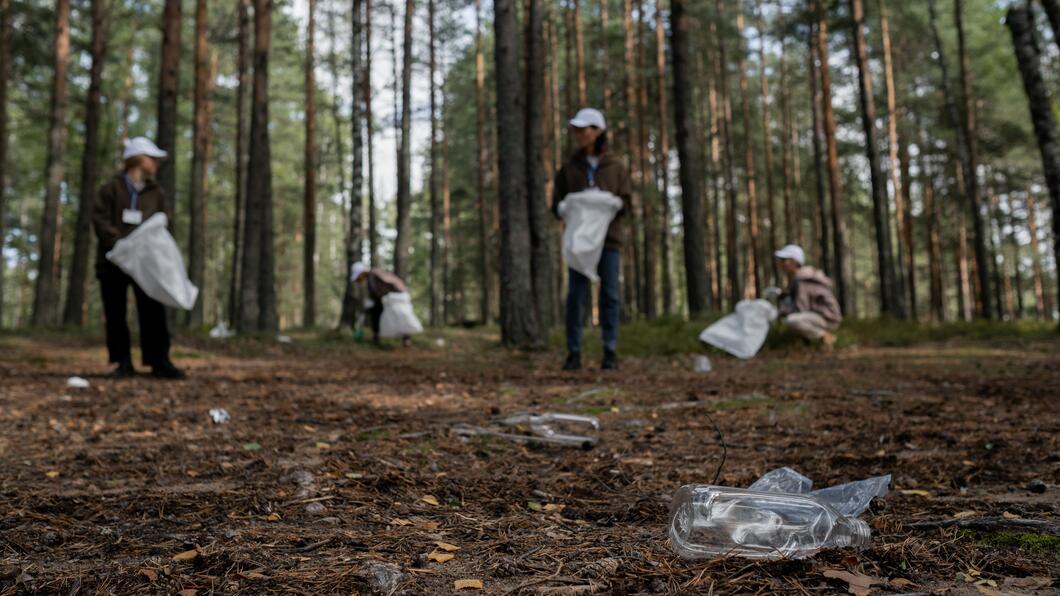 Plastic op de voorgrond. Op de achtergrond zie je mensen in een bos die aan het opruimen zijn. 